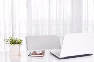 Office table with white laptop, notebook, smartphone, pen and plants placed in the white table with white office chair and white curtain in the background